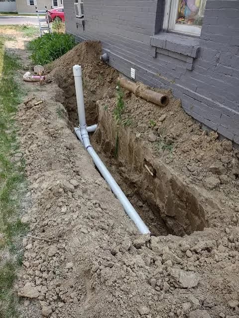 Exterior sewer line trench with new PVC cleanout beside a house foundation